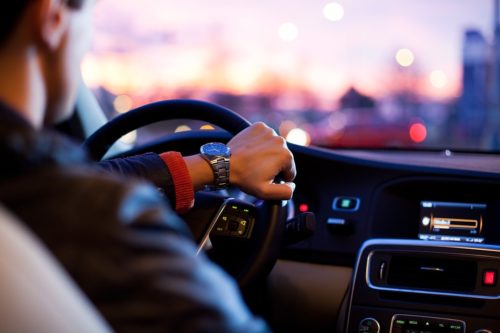 A person wearing a watch drives a car during sunset. The car's interior shows a dashboard with a lit display, and the driver's hand rests on the steering wheel. The view outside the window reveals blurred city lights.