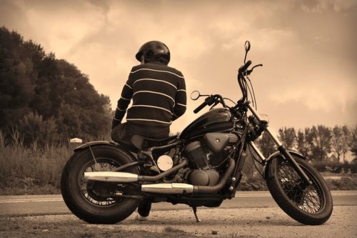 Person wearing a helmet and striped sweater sitting on a black motorcycle, parked on the roadside. The setting is outdoors with trees and an overcast sky in the background, depicted in a sepia tone.