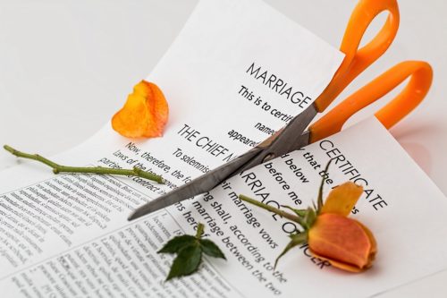 A pair of orange-handled scissors cutting a marriage certificate down the middle, with two orange rose petals and a green stem placed around the document. The image symbolizes separation or divorce.