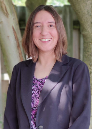 Person with shoulder-length brown hair smiles, standing outdoors in front of a tree. They wear a dark blazer over a purple patterned top. Sunlight filters through the branches, creating dappled light on their outfit.