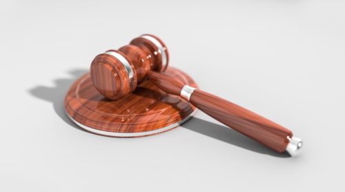 A wooden gavel and sound block on a plain, light gray background, casting a shadow. The gavel is positioned diagonally, showcasing its polished finish and classic design.