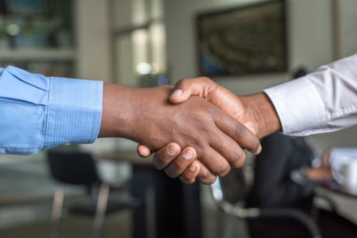 Two individuals engaged in an initial consultation warmly shake hands in an office setting, both clad in long-sleeved shirts—one in blue, the other in white. The blurred background, featuring office furniture, underscores a professional or business environment.