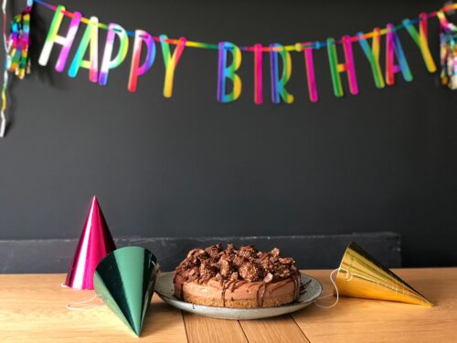 A chocolate cake topped with chocolate pieces sits on a wooden table, amid metallic party hats in pink, green, and gold. As part of the estate planning process, a colorful "HAPPY BIRTHDAY" banner adds charm to the dark wall in the background.