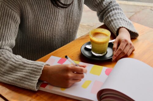 A person in a gray sweater is thoughtfully writing about guardianship in a colorful notebook on a wooden table. Next to them, a glass cup of coffee rests elegantly on a saucer.