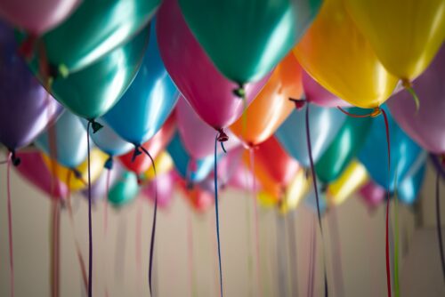 A colorful array of 18 balloons in shades of green, pink, orange, blue, and yellow is shown. They are suspended from the ceiling with ribbons of different colors hanging down. The background is blurred, adding depth to the image.