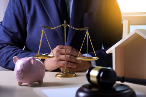 A person in a suit sits at a desk with a scale of justice, a pink piggy bank, a gavel, and a wooden house model, symbolizing law, finance, and the ability to own property in multiple states.