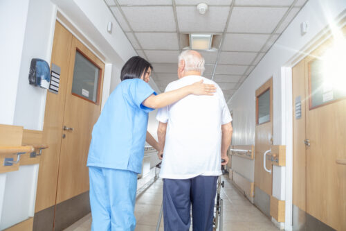 A nurse in blue scrubs assists an elderly man with white hair using a walker in a brightly lit hallway, embodying the role of his healthcare proxy as she gently supports him with her hand on his back while they walk together.
