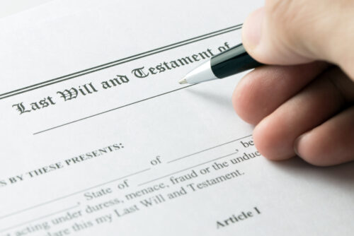 A close-up of a person's hand holding a pen, preparing to sign a document titled "Last Will and Testament." The paper has blank spaces for details to be filled in.