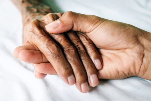 A younger hand gently holding an older hand, both resting on a white surface, conveying comfort, care, and support between generations—illustrating the role of a conservator in safeguarding an elder’s estate.