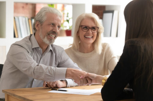 Smiling older couple sits at a table, shaking hands with a woman across from them, appearing happy and satisfied during a meeting or agreement in a bright office setting.