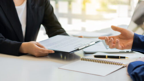 Two people in business attire sit at a desk; one holds a clipboard with documents, while the other gestures with a hand. An open notebook, pen, and laptop are also on the desk.