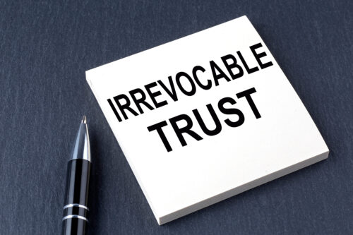 A black pen rests beside a white notepad labeled "IRREVOCABLE TRUST" in bold black letters, highlighting the importance of an irrevocable trust against a sleek, dark background.