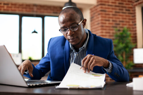 A man wearing glasses and a blue blazer sits at a desk, looking at documents and working on a laptop in an office with brick walls and large windows as he reviews his year-end estate plan.