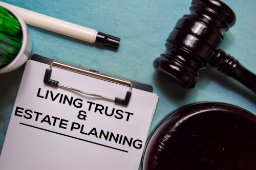 A clipboard with a paper titled "LIVING TRUST & ESTATE PLANNING" sits next to a pen, a plant, and a judge’s gavel on a blue surface, representing legal and financial planning for a living trust or revocable trust.