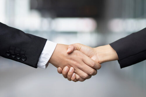 Close-up of two people in business attire shaking hands, symbolizing agreement or partnership, with a blurred indoor background.