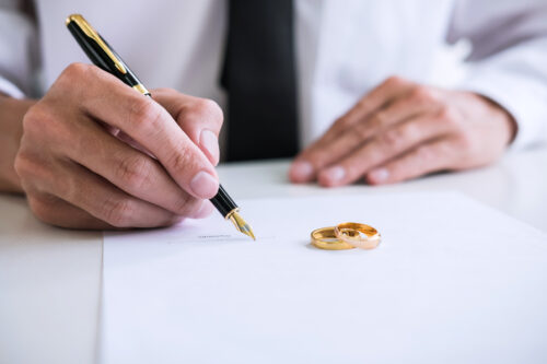 A person in a white shirt signs a document with a fountain pen. Two gold wedding rings rest on the paper, suggesting the signing of a divorce or marriage-related agreement.