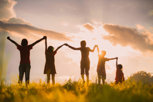 Five children hold hands and raise their arms while standing in a grassy field at sunset, their silhouettes outlined against the bright sky and clouds.